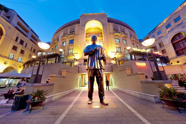 A large bronze statue of a man in a loose shirt, his arms poised in dance. He stands at the lit-up entrance to shops.