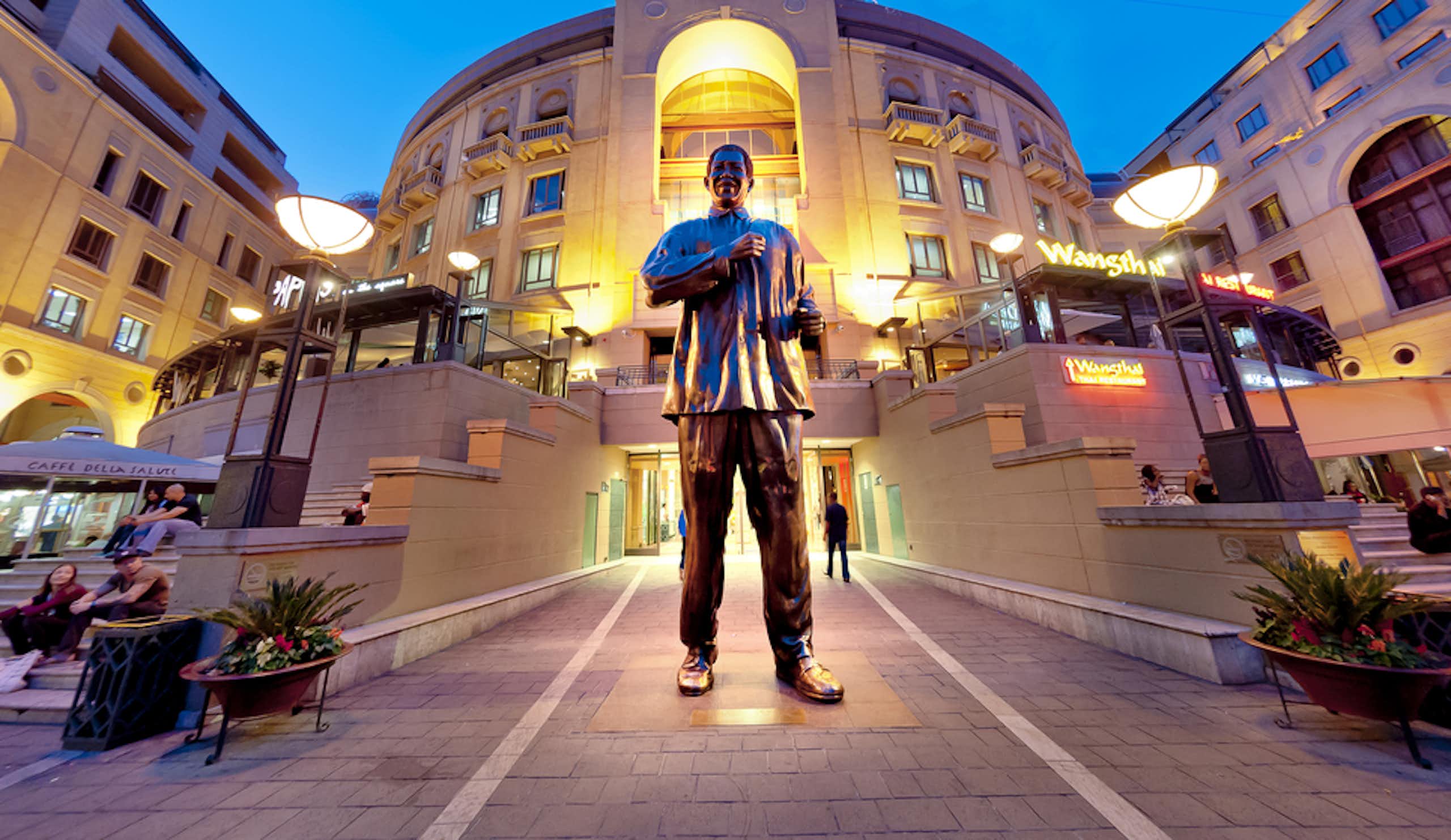 A large bronze statue of a man in a loose shirt, his arms poised in dance. He stands at the lit-up entrance to shops.