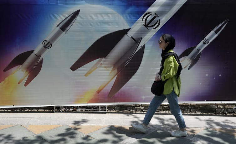 A woman in jeans and a head scarf walks past a banner showing missiles.