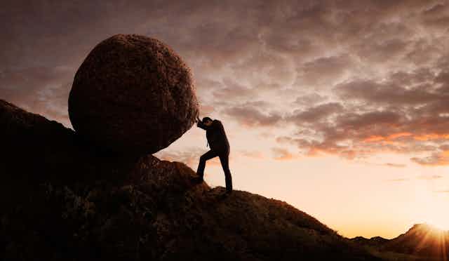 A man in a business suit pushing a boulder up a mountain