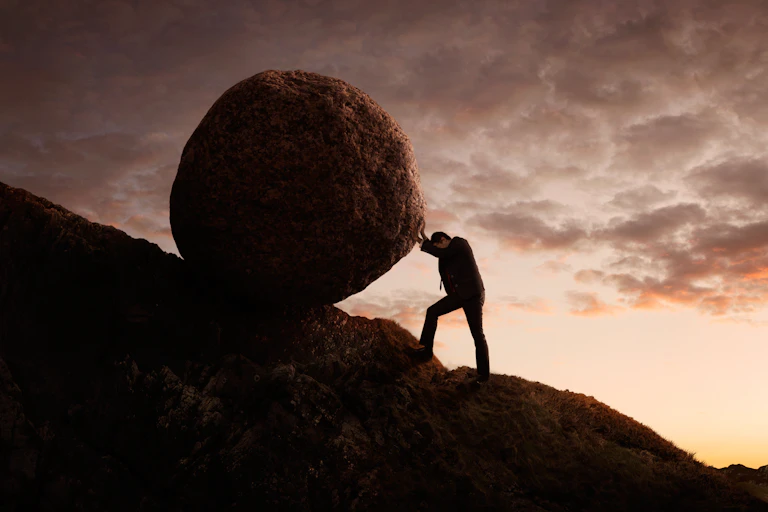 A man in a business suit pushing a boulder up a mountain