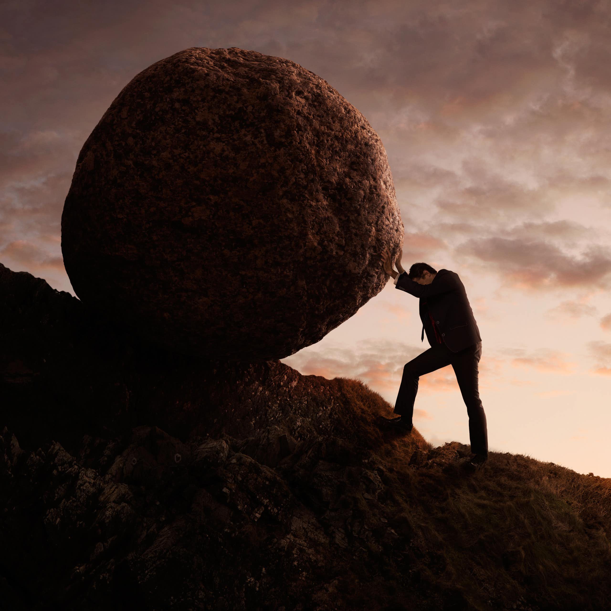 A man in a business suit pushing a boulder up a mountain