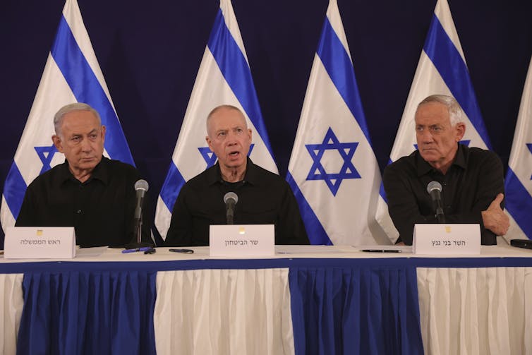 Three older men sit in a row at a table with blue and white flags behind them.