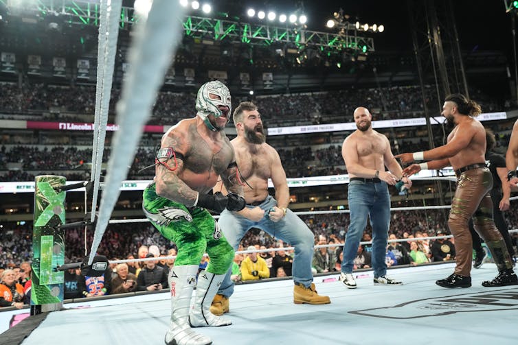 Four shirtless men, one wearing green leggings and a face mask, stand in a wrestling ring in a crowded stadium