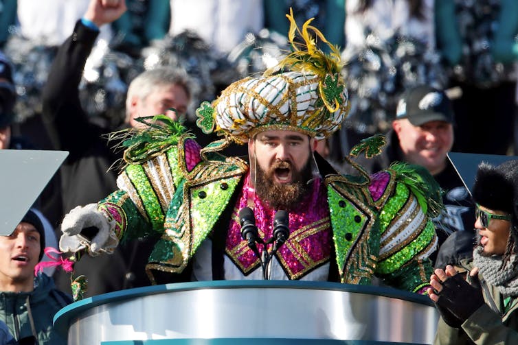 A tall bearded man wearing an elaborate pink and green costume and a gold hat speaks at a podium