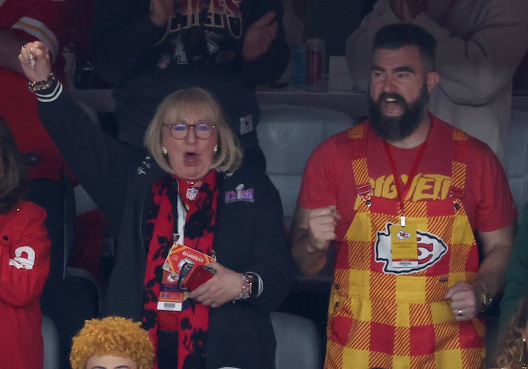 A woman cheers with her fist in the air and stands beside a bearded man wearing red and yellow checkered overalls