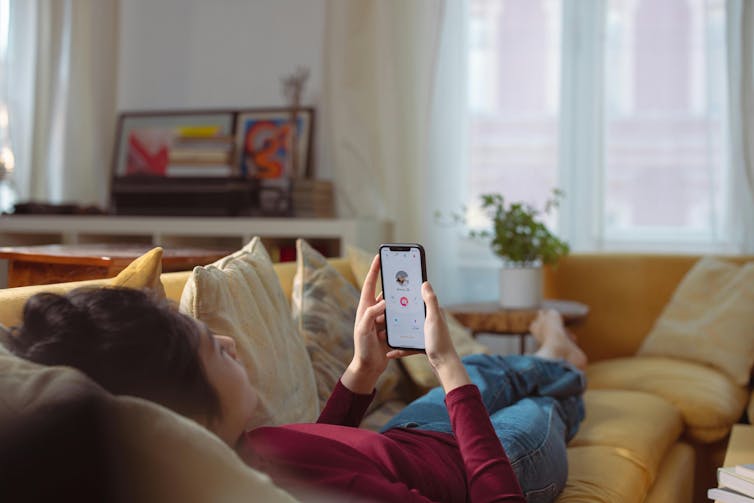 A person lies on a sofa with a phone and a dating app can be seen on the screen