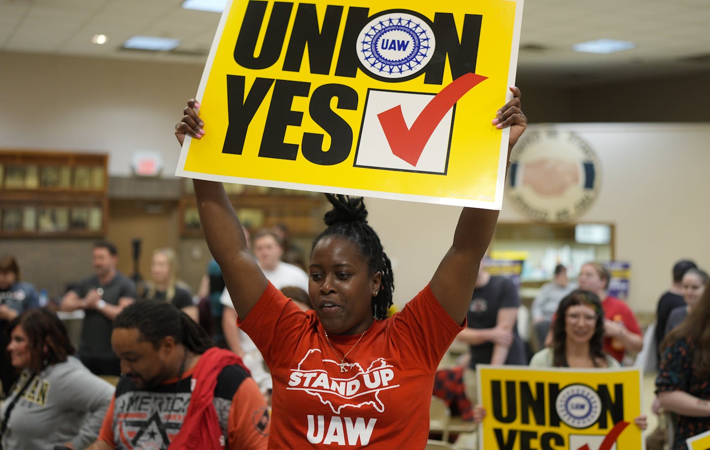 A woman in a red t-shirt saying 'stand up UAW' holds a yellow sign aloft that says 'UnionYes' with a checkmark.
