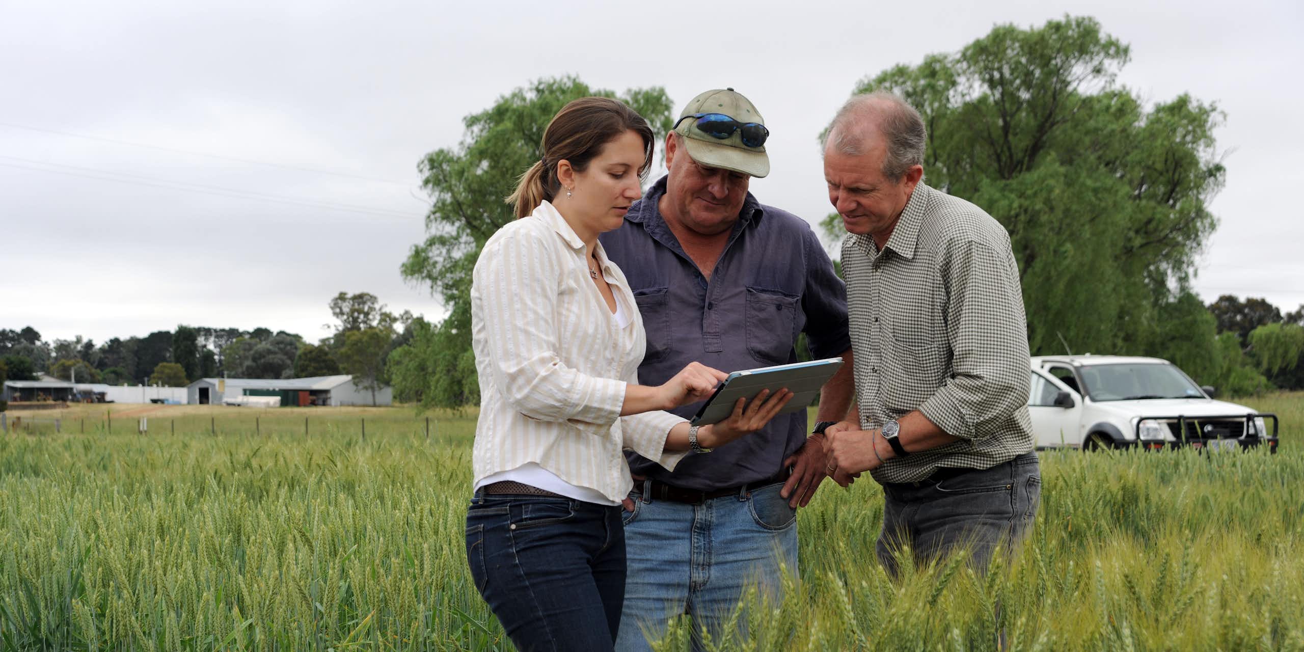Three people gather around an iPad or table in a field, with farm vehicles in the background