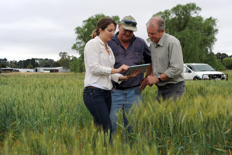 Three people gather around an iPad or table in a field, with farm vehicles in the background