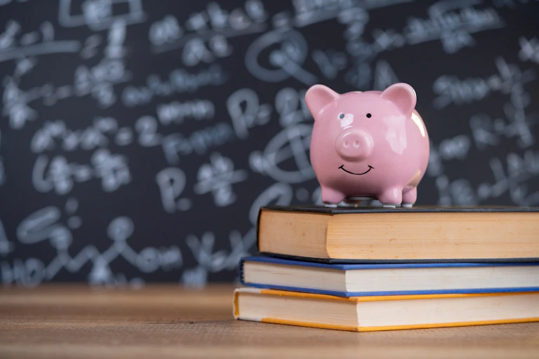 A pink piggy bank on a pile of books, with a blackboard in the background
