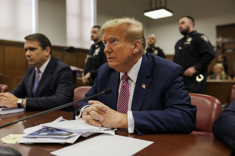 Two midle-aged white men dressed in business suits sit at a table in front of several police officers.