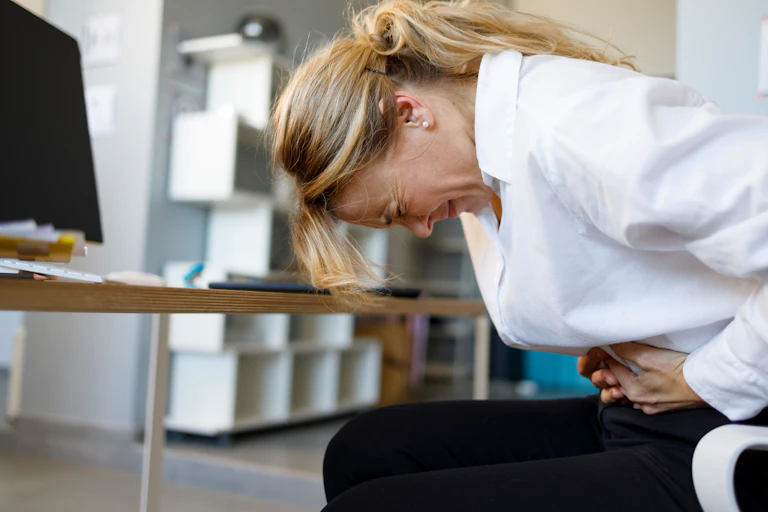 Woman seated at a desk holding her stomach suggesting she is unwell.