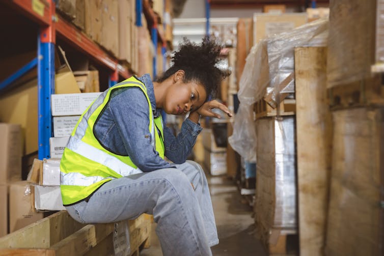 Woman warehouse worker in high visibility best sitting down, holding her head.