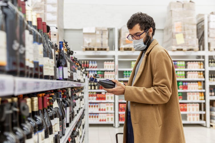 Man shops for alcohol in a store. He wears a large coat and a face mask.