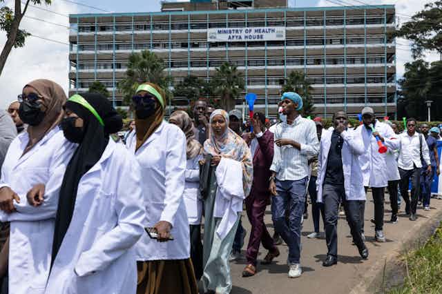 Women and men walking on a street wearing white coats, a large building behind them.