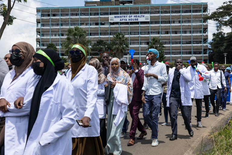 Women and men walking on a street wearing white coats, a large building behind them.