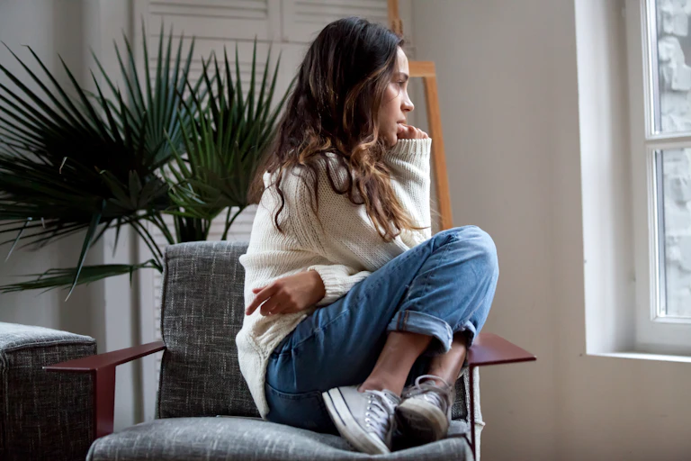 A young woman sitting on a chair, knees bent, looking pensive