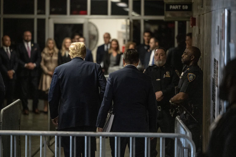 A man with golden hair is walking into a courtroom surrounded by police officers and men and women dressed in business attire.