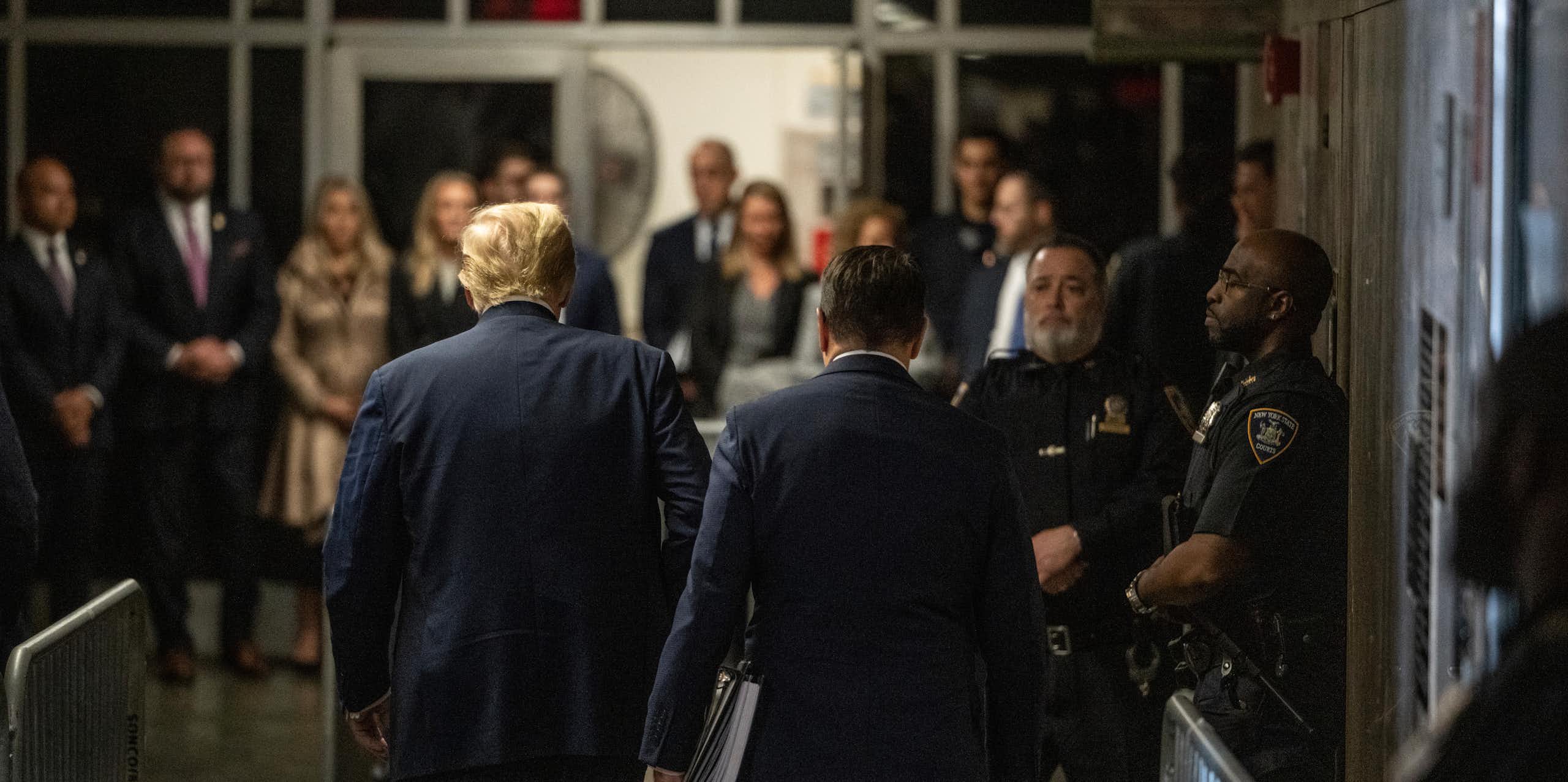 A man with golden hair is walking into a courtroom surrounded by police officers and men and women dressed in business attire. 