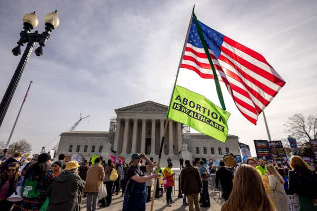 A person holds an American flag and a sign that says 'Abortion is healthcare,' outside the Supreme Court building, surrounded by other people behind barricades. 