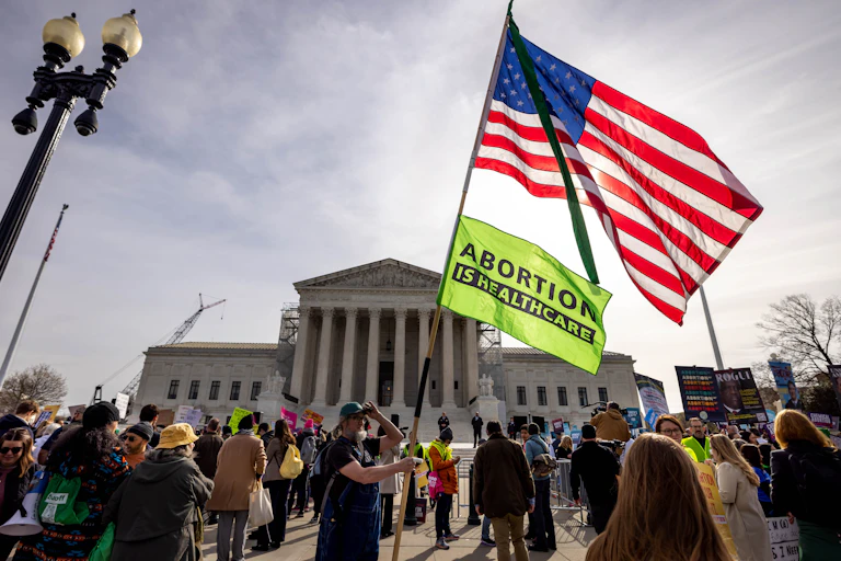 A person holds an American flag and a sign that says 'Abortion is healthcare,' outside the Supreme Court building, surrounded by other people behind barricades.