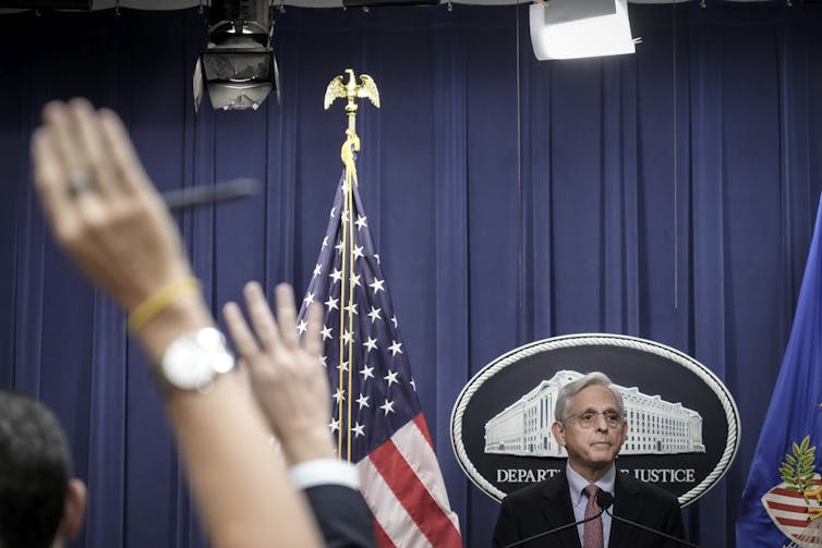 A white man with gray hair stands at a podium in front of a White House seal, staring over people with their hands raised in the air.