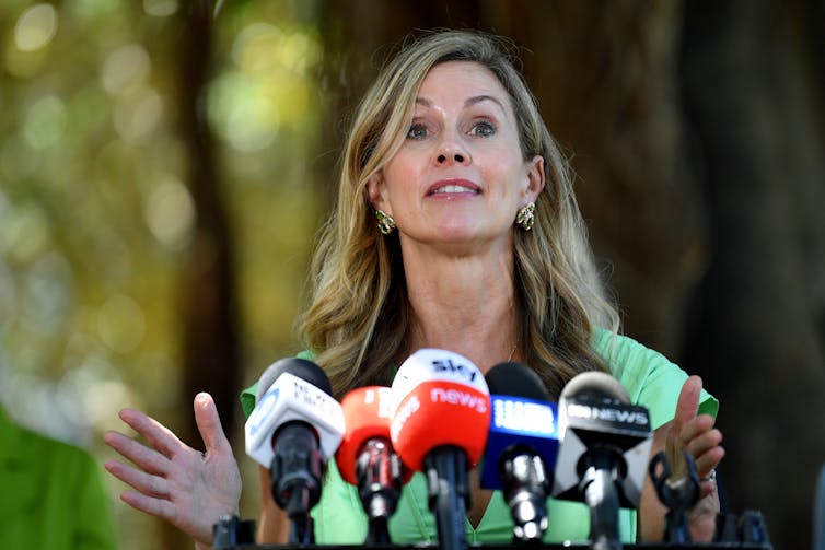 A blonde woman with an energetic expression stands in front of an array of microphones.