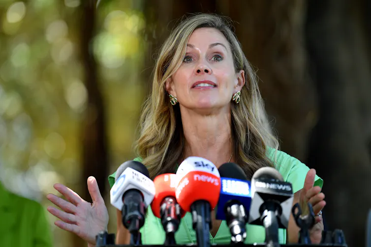 A blonde woman with an energetic expression stands in front of an array of microphones.
