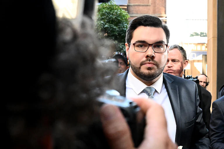 A man in glasses and a suit and tie walks outside surrounded by media cameras