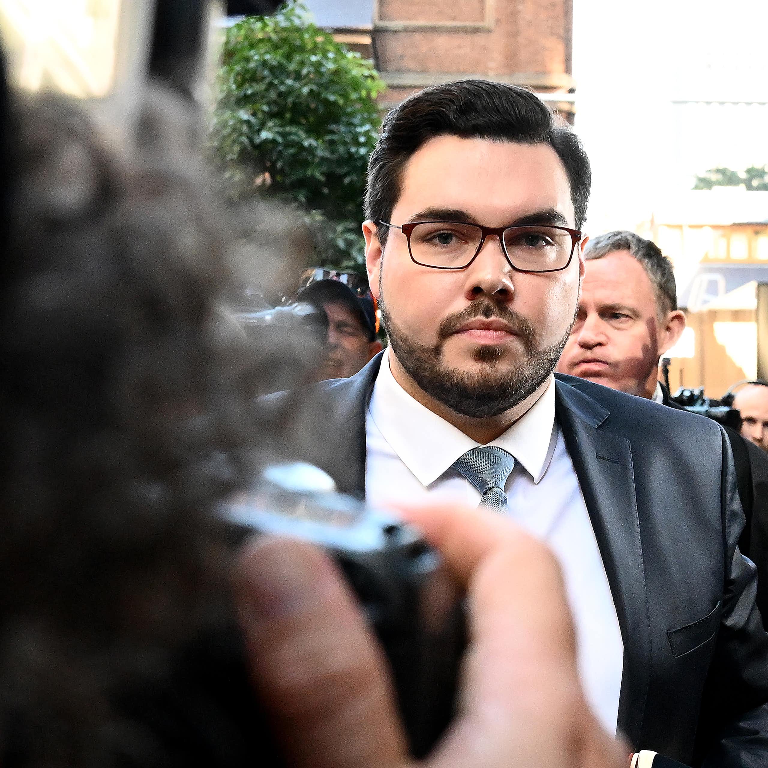 A man in glasses and a suit and tie walks outside surrounded by media cameras