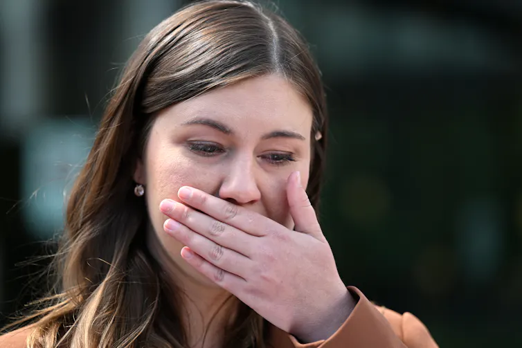 A woman with brown hair covers her mouth with the hand as she tears up.