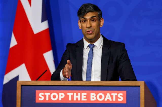 Rishi Sunak speaking and gesturing decisively at a podium that reads Stop the Boats