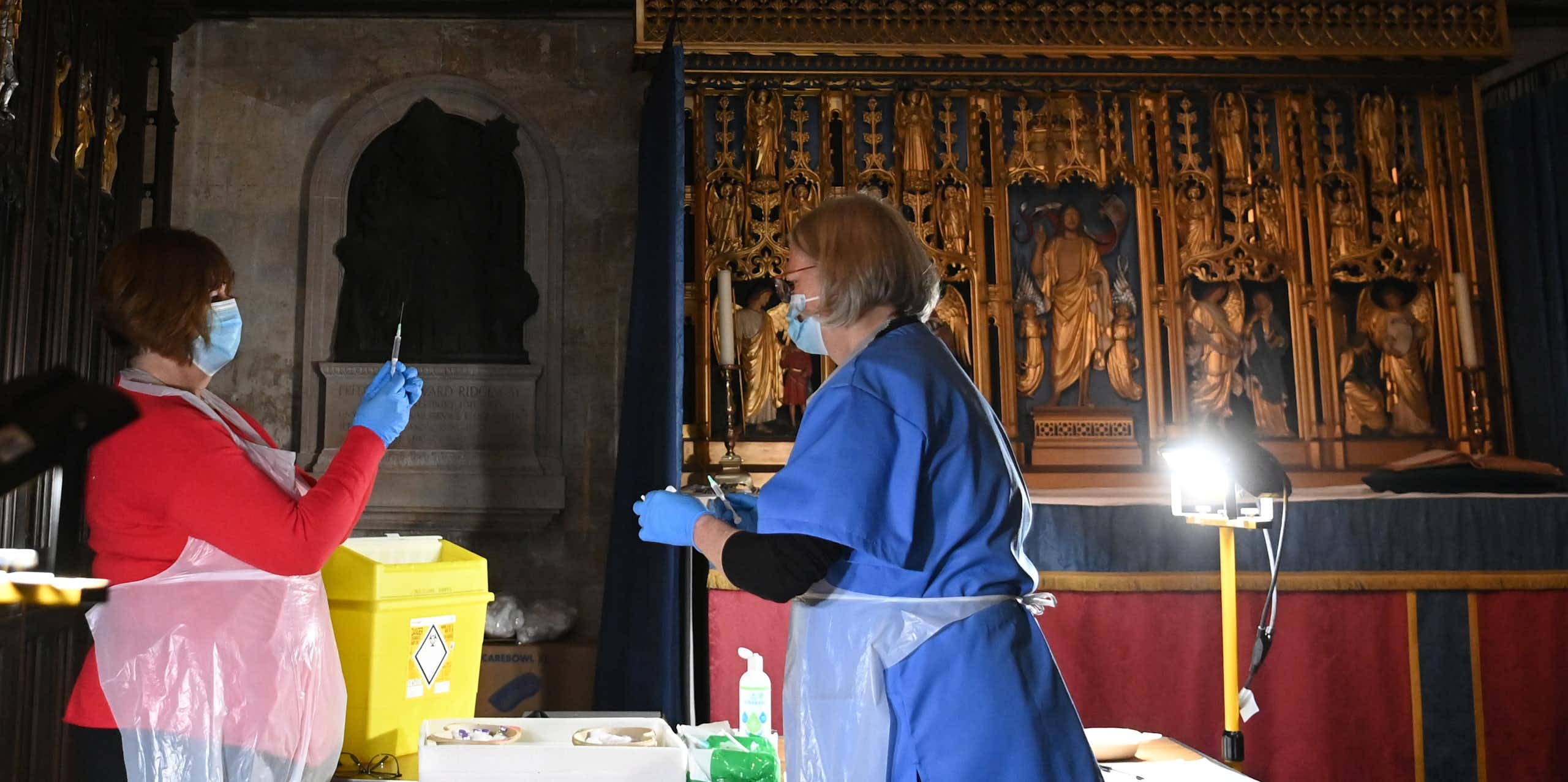 Two women in PPE prepare vaccines inside a church. 