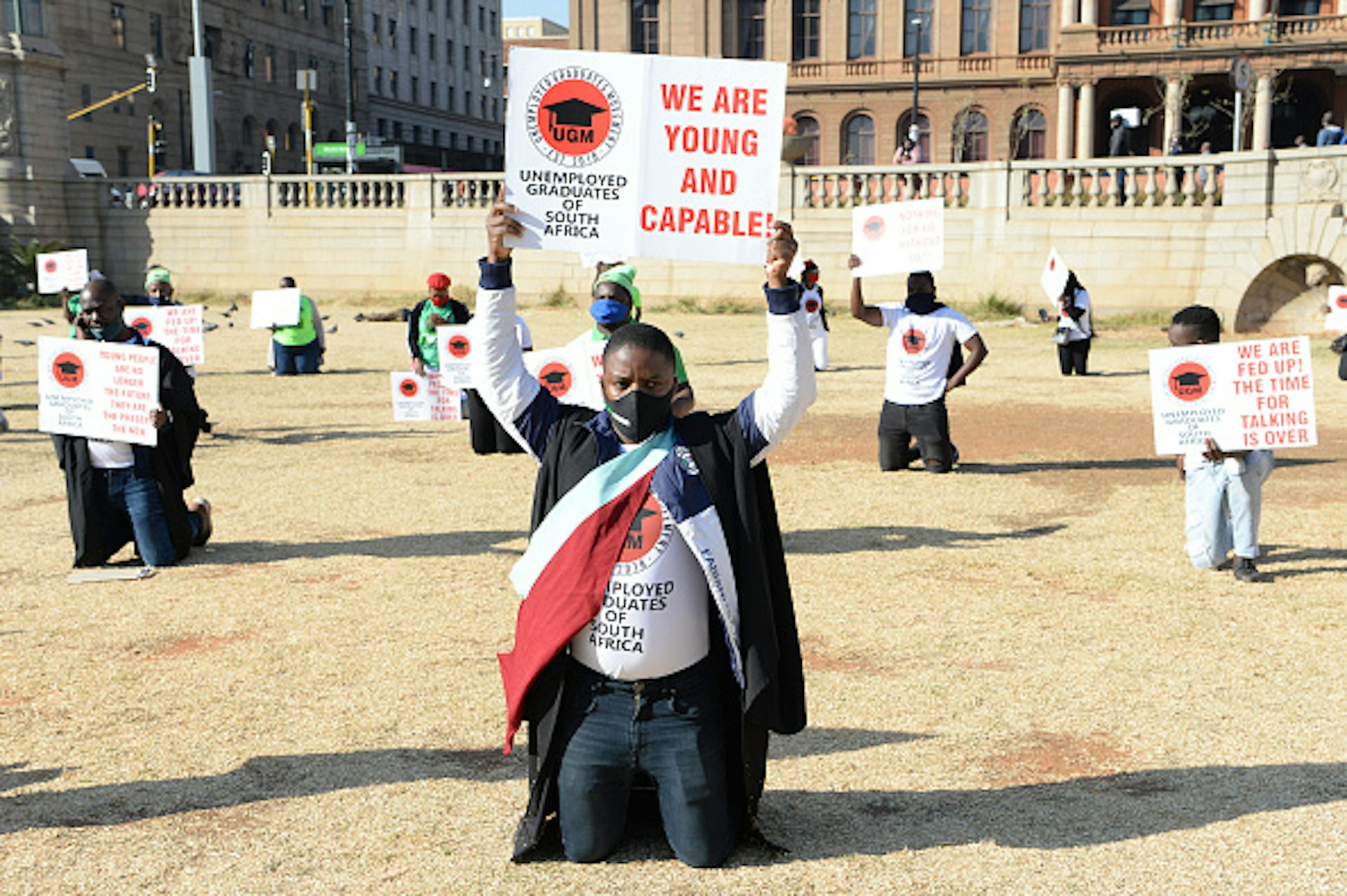 Two young men wearing graduation gowns lead protesters carrying placards saying 'We are young and capable'.