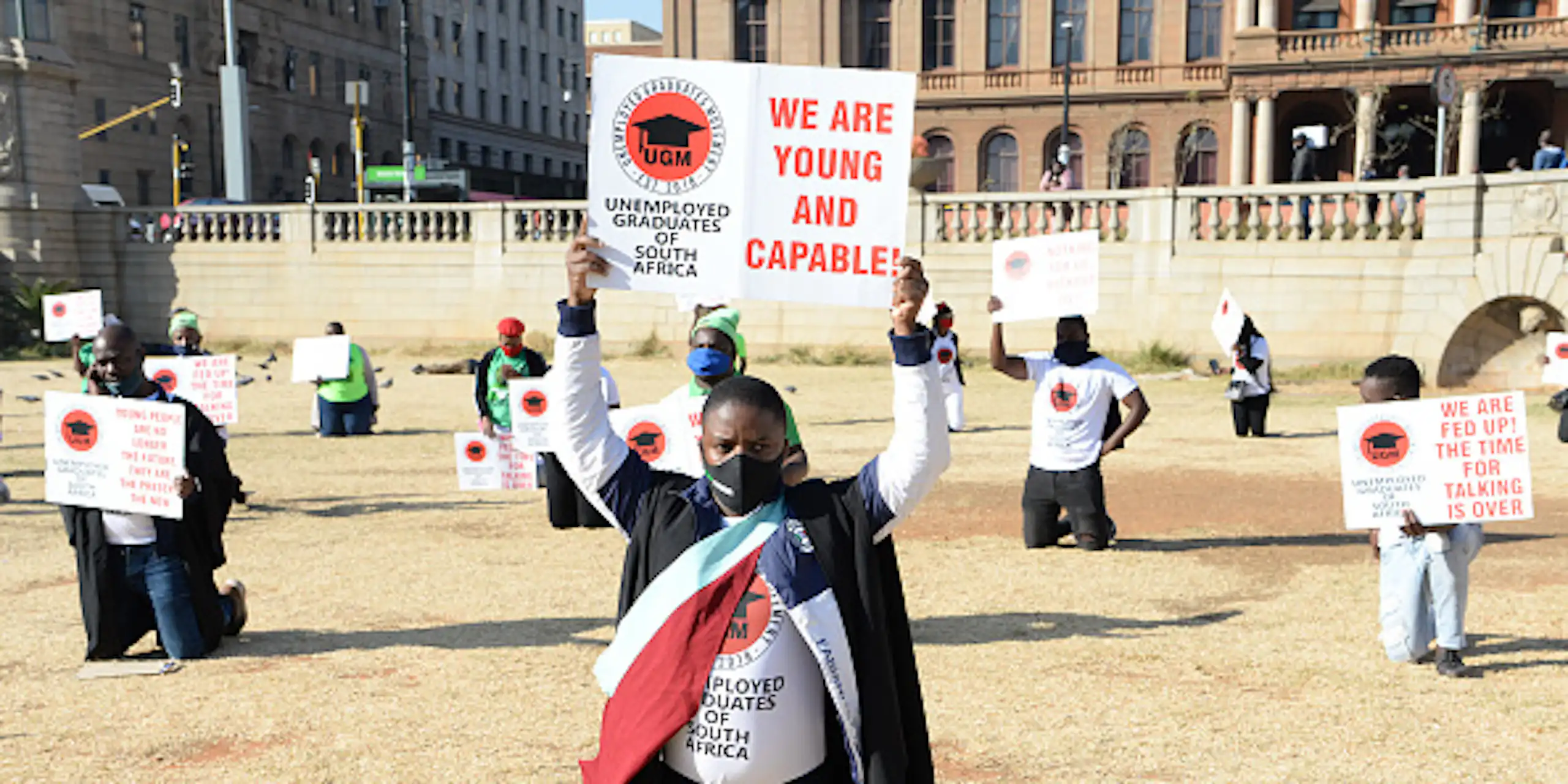 Two young men wearing graduation gowns lead protesters carrying placards saying 'We are young and capable'.