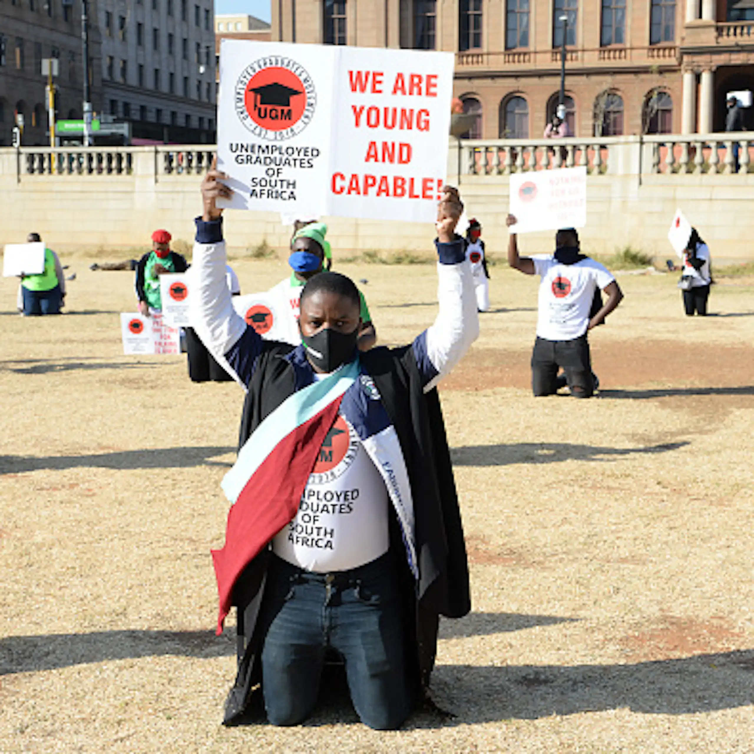 Two young men wearing graduation gowns lead protesters carrying placards saying 'We are young and capable'.