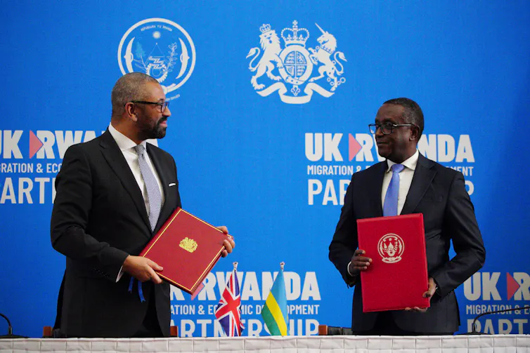 Home secretary James Cleverly and Rwandan Minister of Foreign Affairs Vincent Biruta standing at a table each holding a red folder after signing a new treaty