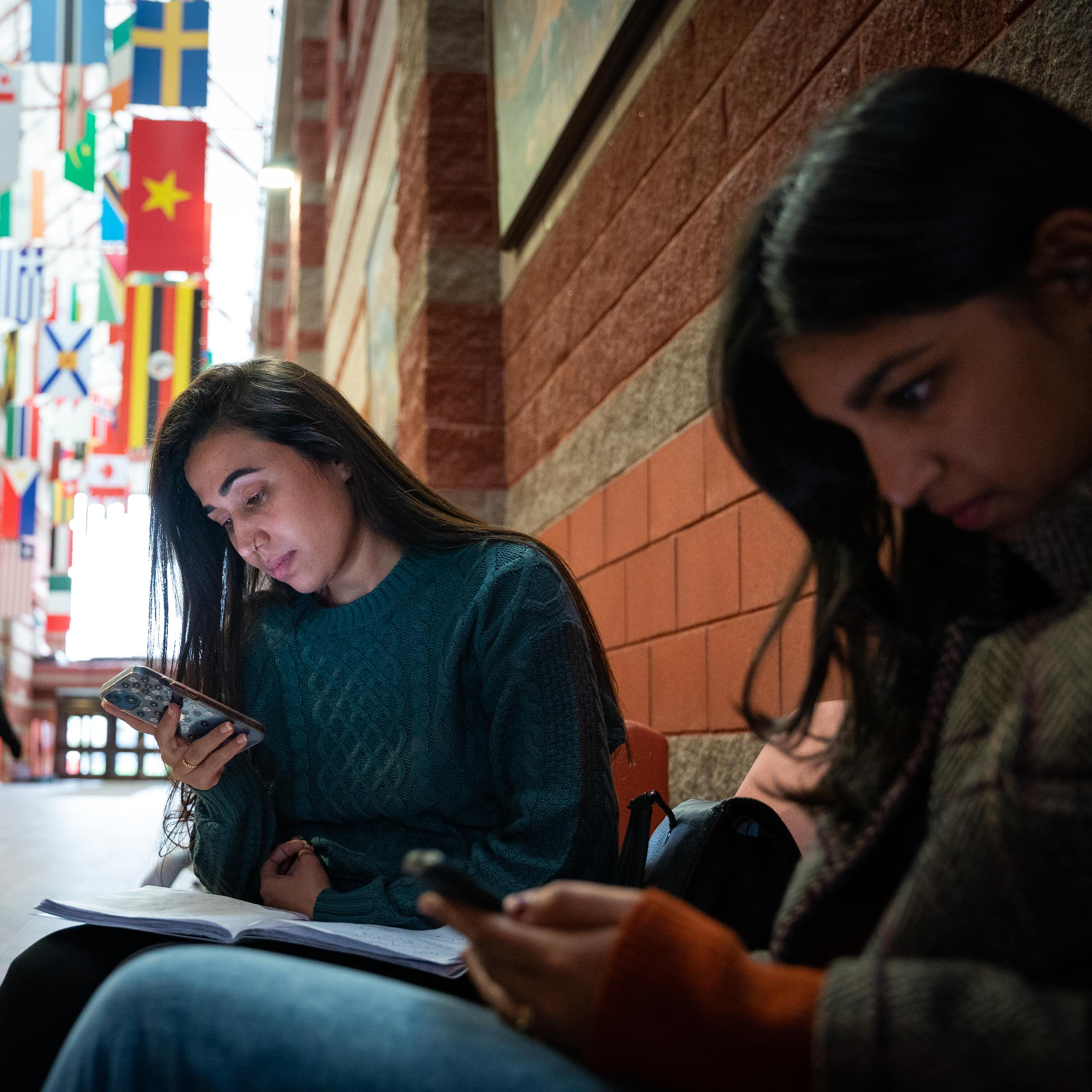 Two dark-haired girls look at their phones as they sit in a hall at a university. One has a notebook on her lap.