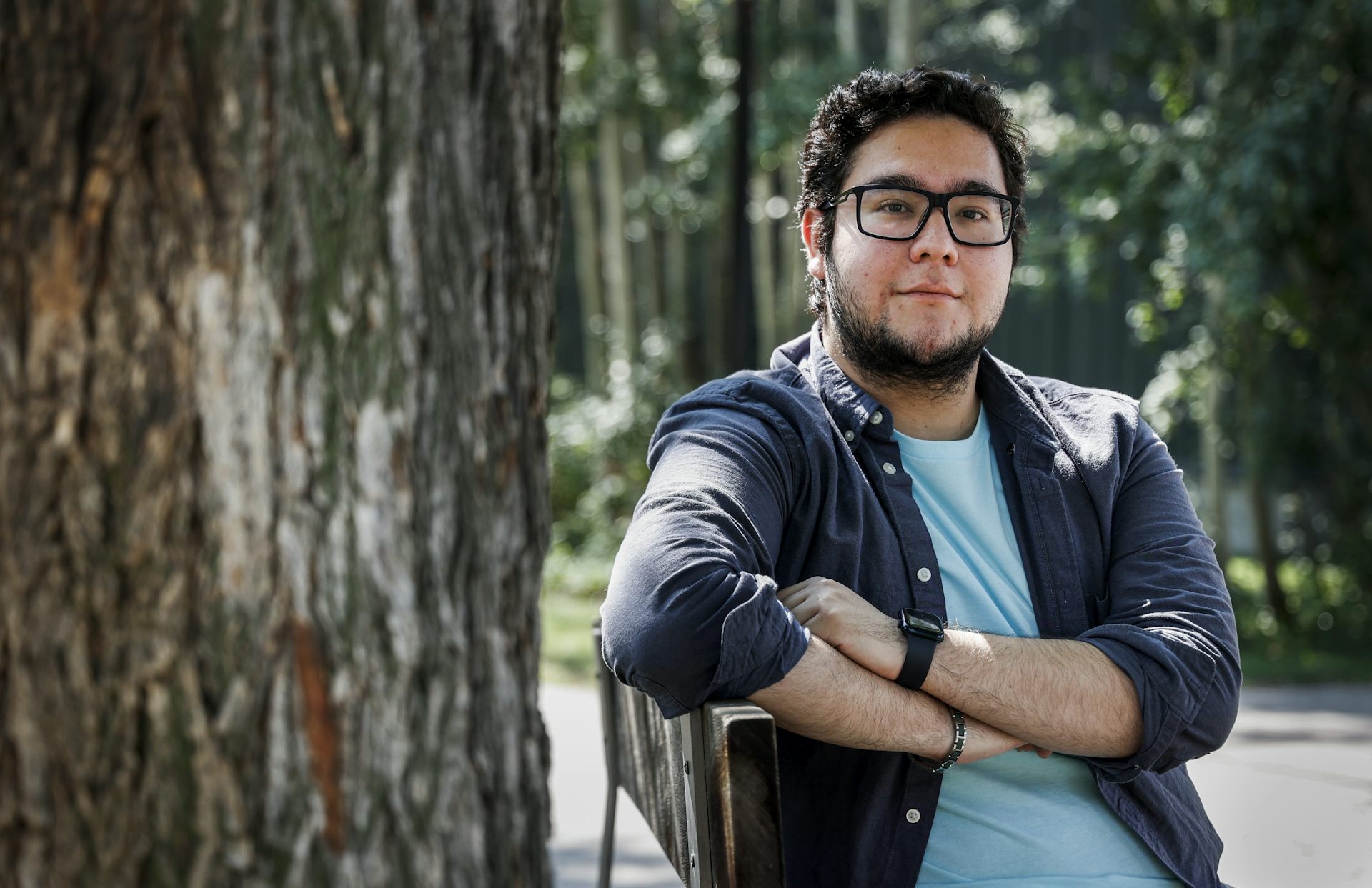 A young man with a beard and glasses sits on a bench next to a tree with his arms crossed.