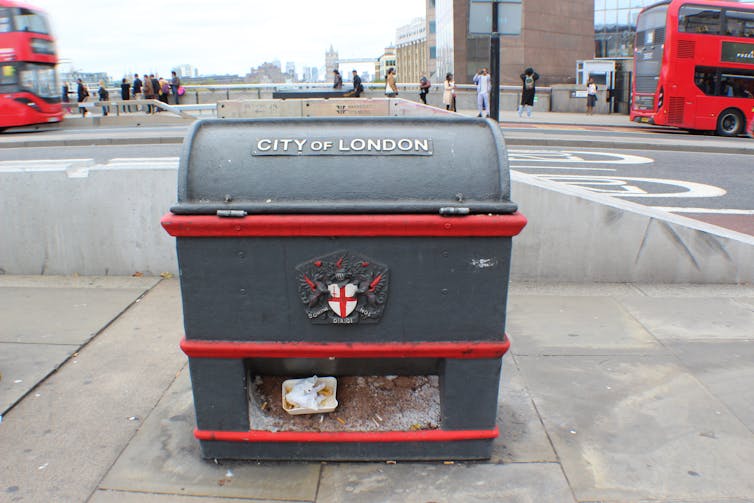 urband London roadside, pavement with black and red rectangular box with small white plastic rubbish in a crevice underneath, red buses in distant background
