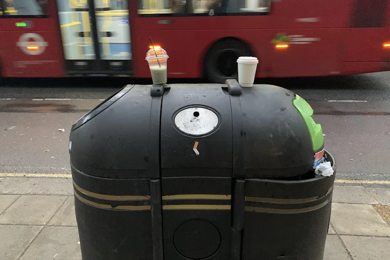 London street, big black bin in foreground, two white plastic cups sitting littered on top, blurry red bus in background