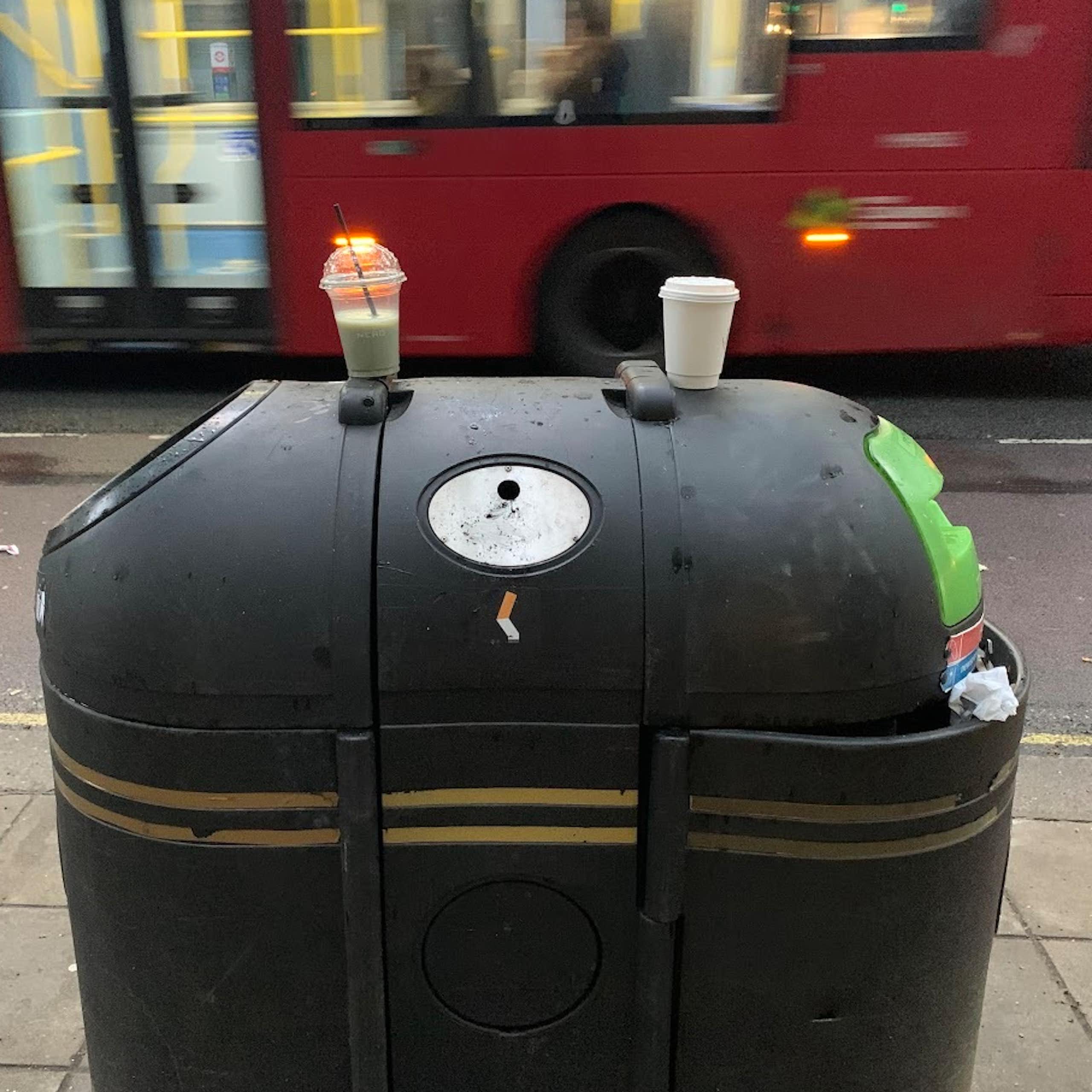 London street, big black bin in foreground, two white plastic cups sitting littered on top, blurry red bus in background