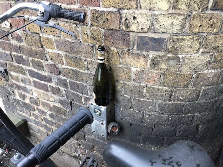 brick wall, part of a black bicycle in foreground, open green wine bottle resting on a small ledge against bricks