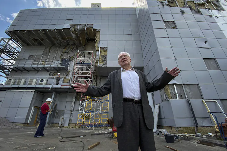 A man in a suit stands in front of a damaged building.