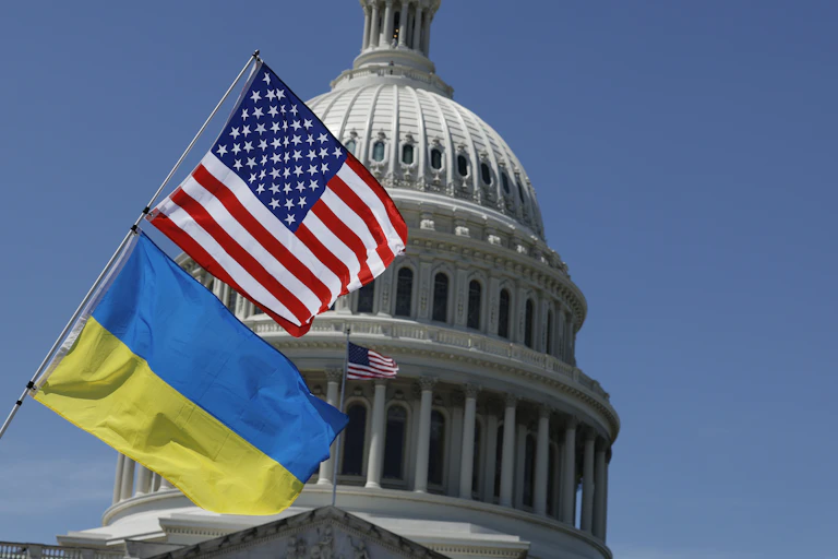 A blue and yellow flag rests on a poll below an American flag, in front of the top of the Capitol building dome. There is a bright blue sky behind the building.