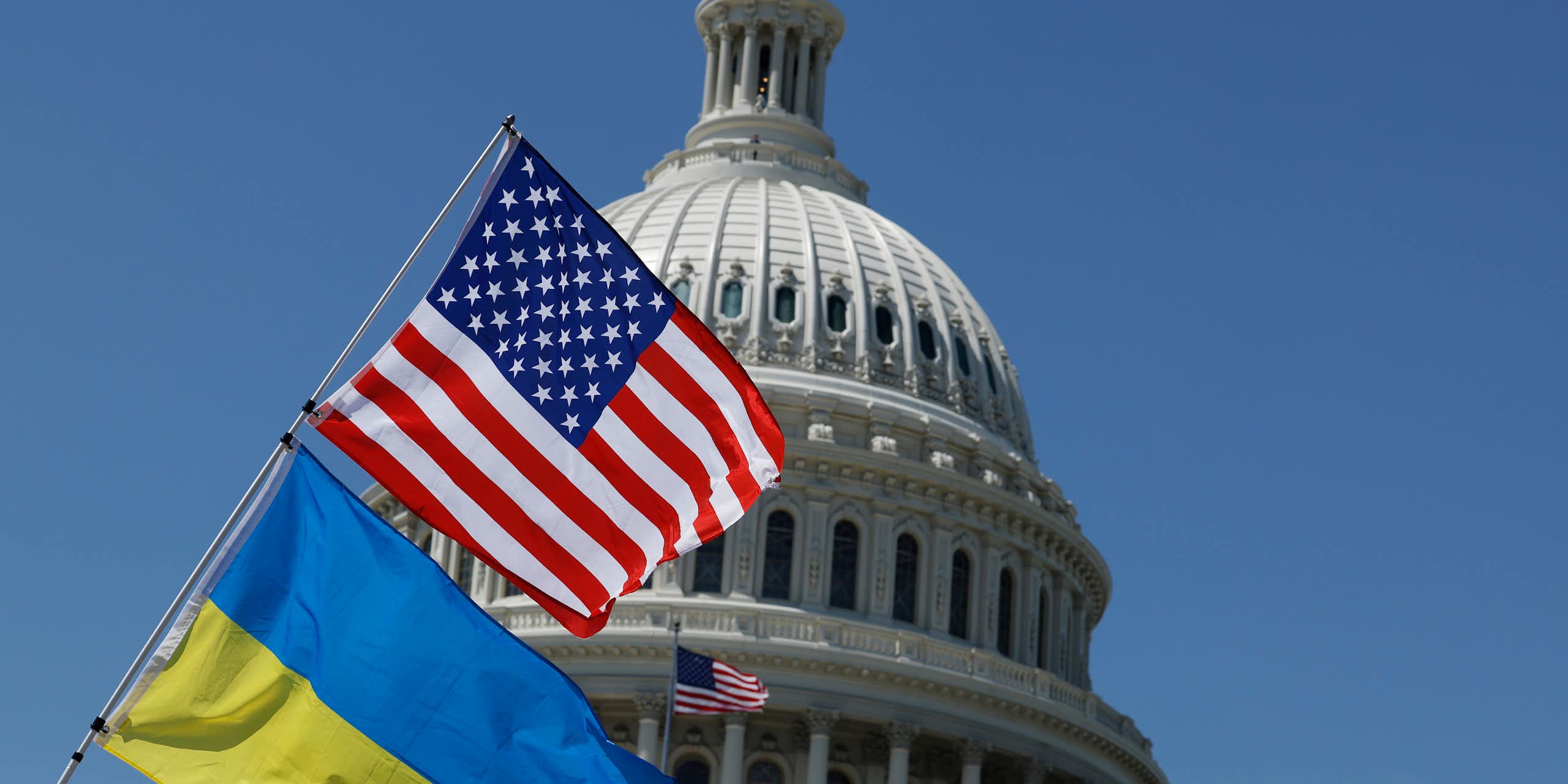 A blue and yellow flag rests on a poll below an American flag, in front of the top of the Capitol building dome. There is a bright blue sky behind the building.