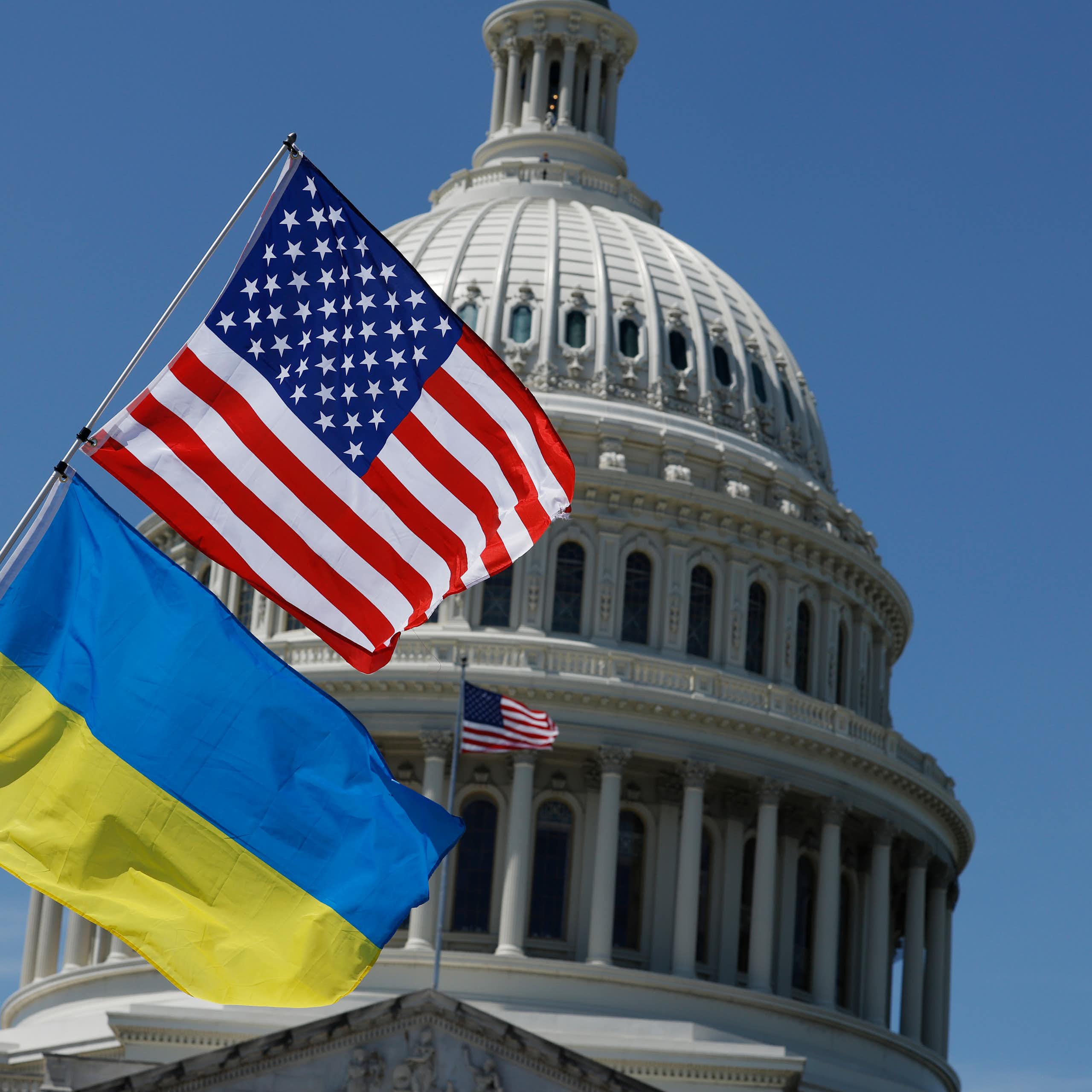 A blue and yellow flag rests on a poll below an American flag, in front of the top of the Capitol building dome. There is a bright blue sky behind the building.