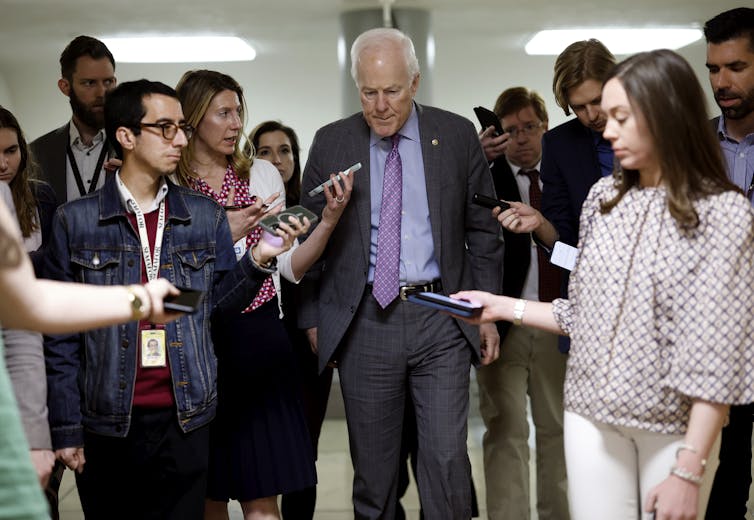 A white man with white hair and a grey suit and purple tie walks surrounded by people who extend cell phones and tape recorders in his direction.