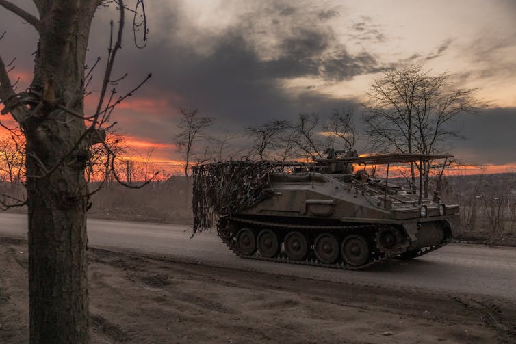 A tank is seen driving on an empty road, with a smoky sky and an orange sunset behind it.